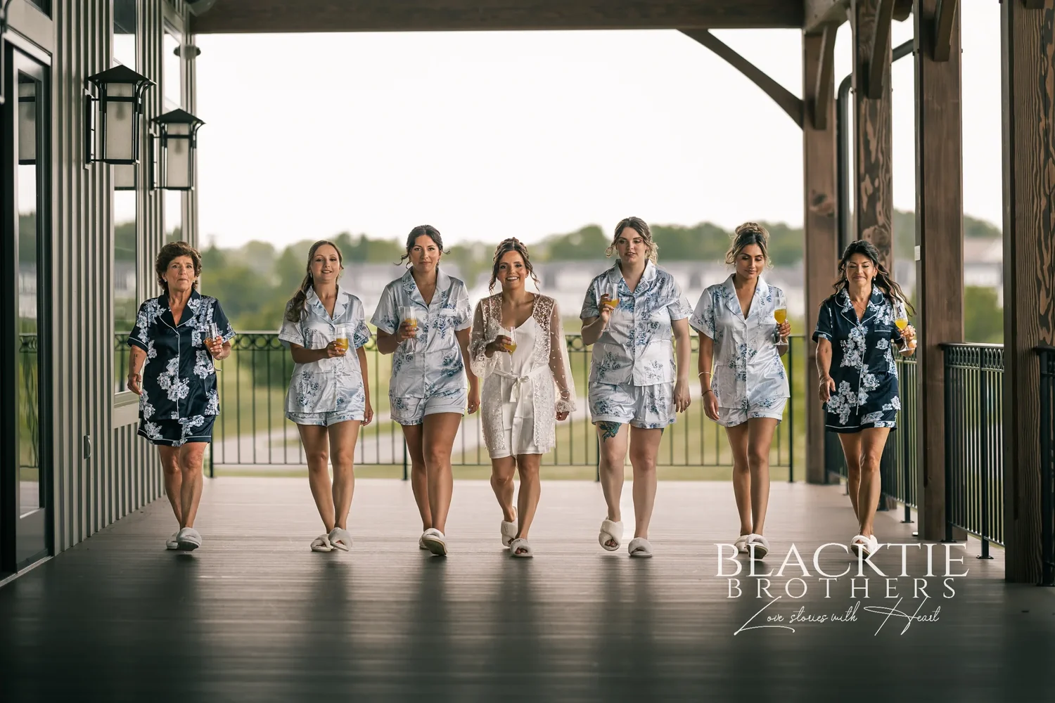 Taylor in a white robe and her bridesmaids in blue floral pajamas walking toward the camera along the covered timber porch, mimosas in hand, the golf course behind them