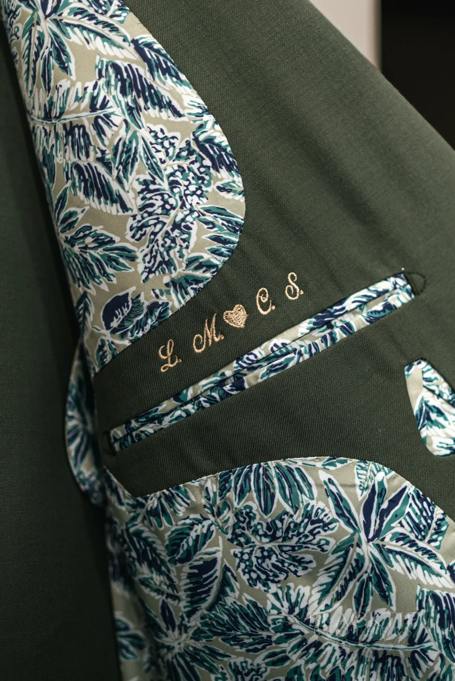 The inside of a sage-green suit jacket with a patterned lining, embroidered with the couple’s initials and a heart