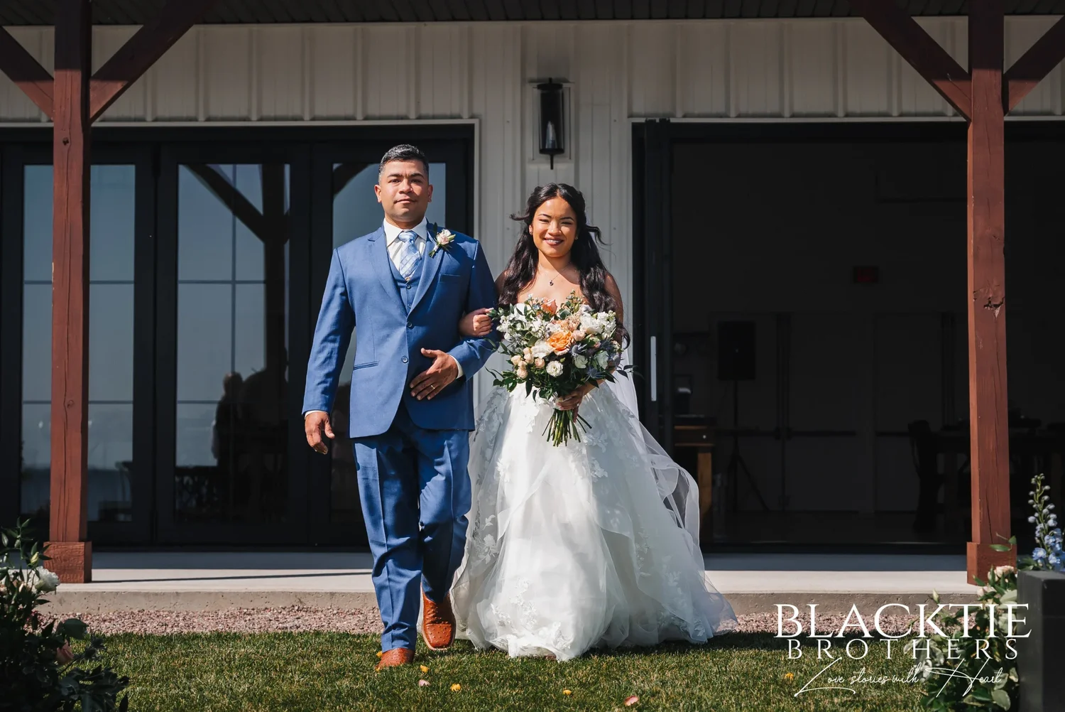 Carolina holding a bouquet of peach and white flowers as she walks out of the event center on the arm of a man in a blue suit