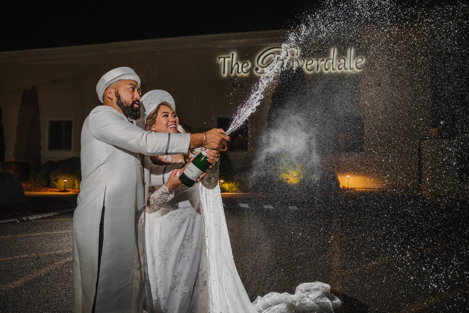 Alice and Mitchell, changed into white formal outfits with matching headpieces, spraying champagne in front of the lit Riverdale sign at night