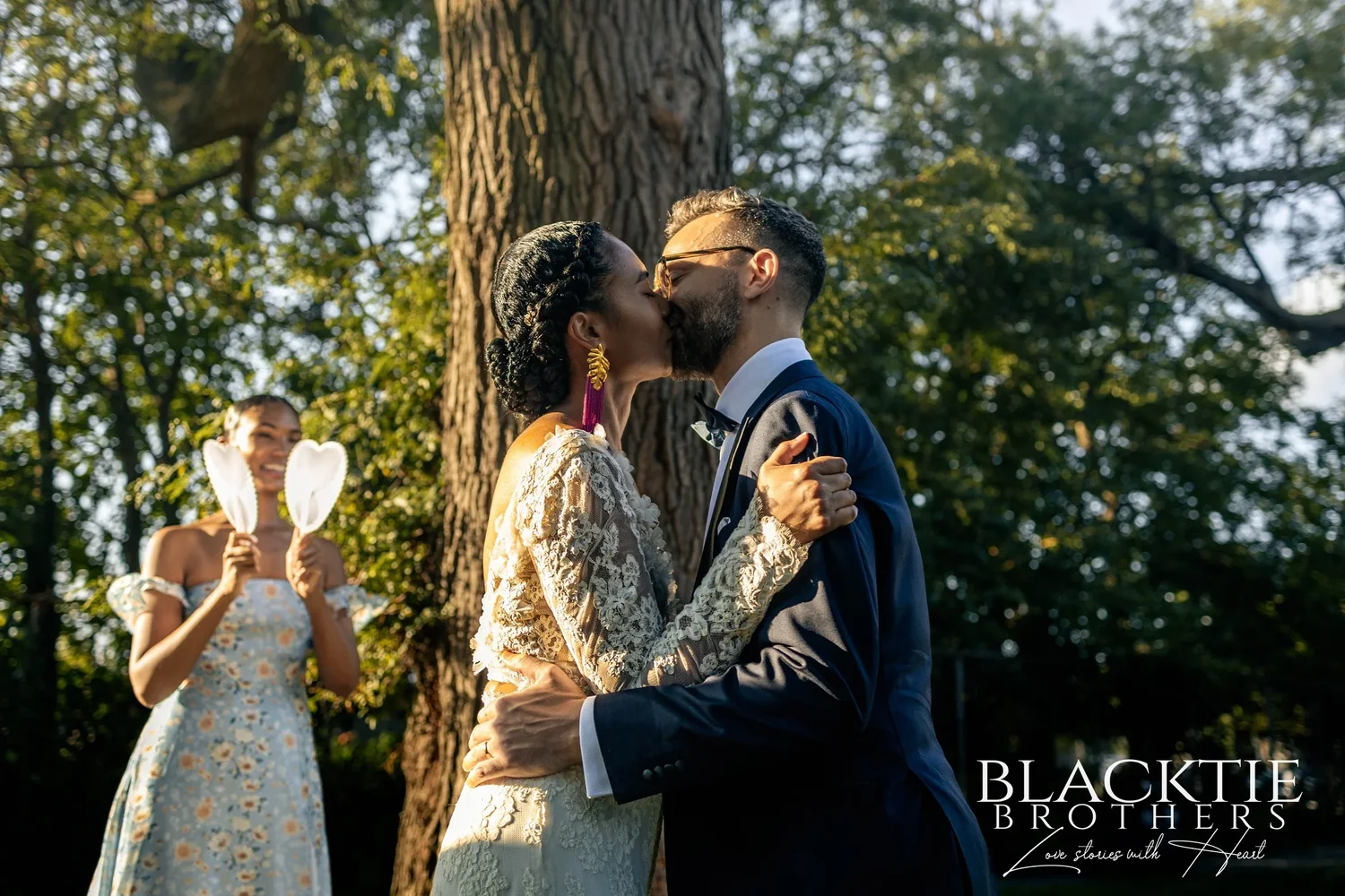 Ciara and Drew's first kiss under the tree as a member of the wedding party cheers behind them holding two white fans