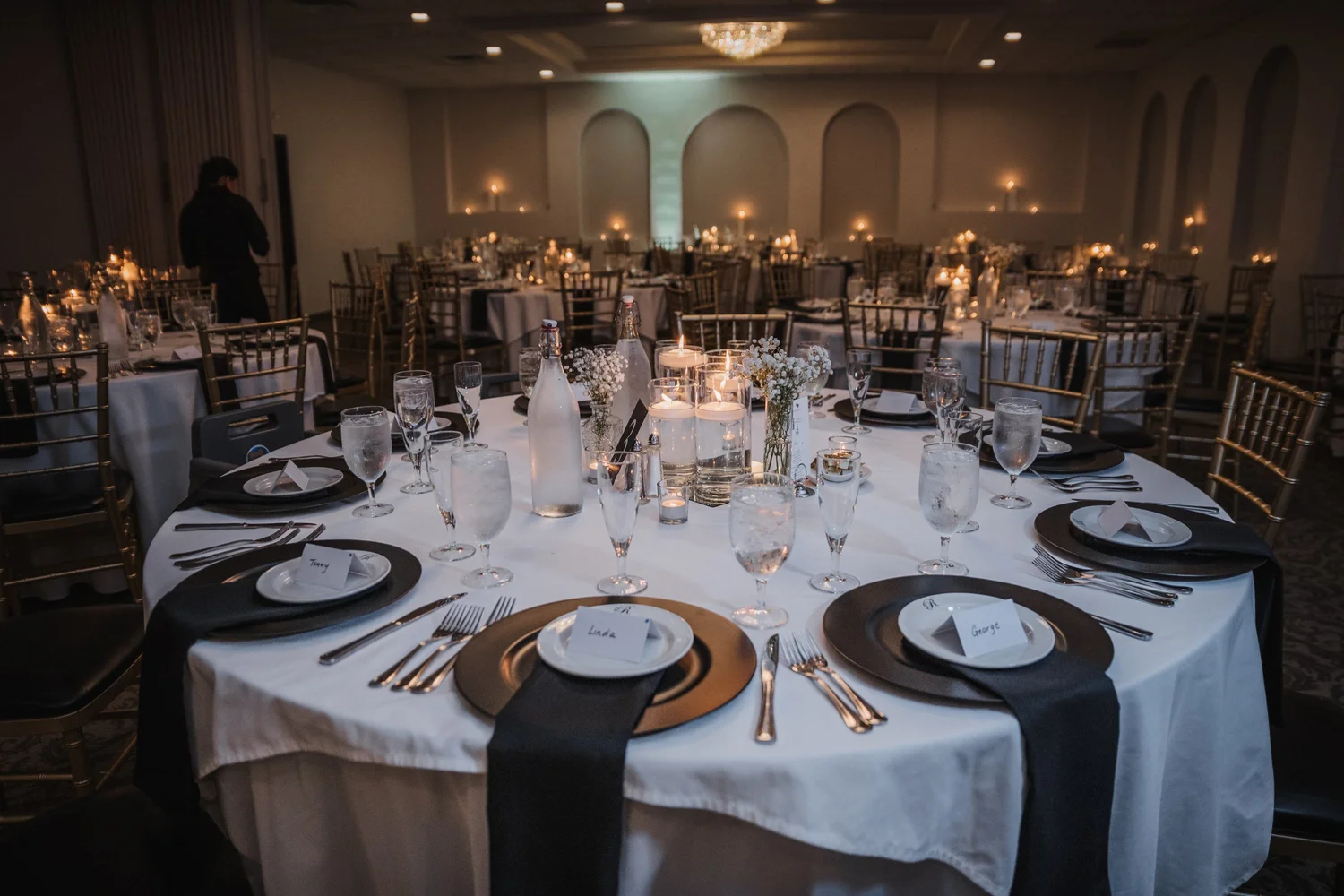 Reception tables at The Riverdale set with white cloths, black napkins and gold chargers, candles glowing across the room