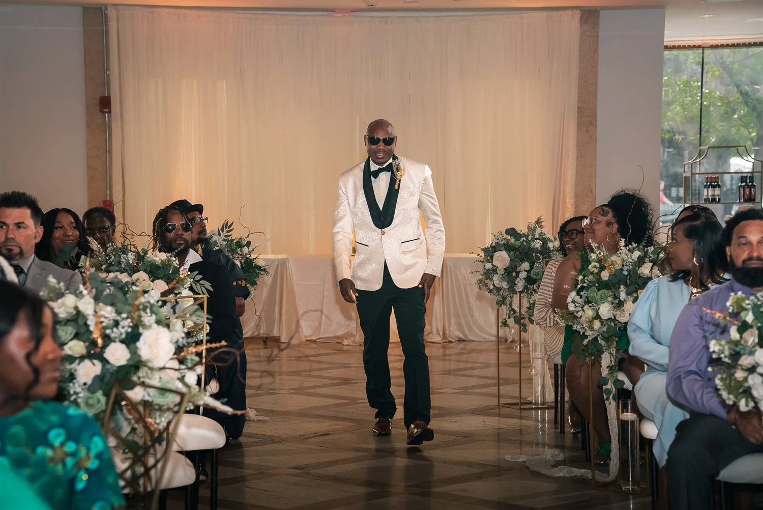 David walks between rows of wedding guests wearing an ivory jacket with dark green lapels.