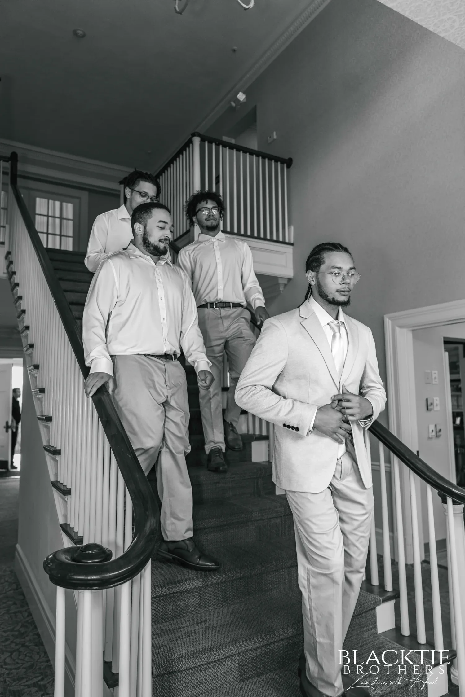 Black-and-white photograph of a groom in a light suit buttoning his jacket as he walks down the mansion staircase at Traditions at the Glen, his groomsmen following behind