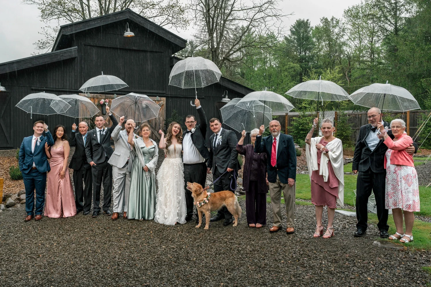 Lindsey and Noah with family and friends under clear umbrellas in front of the Black Barn on a rainy day, a golden retriever in the front row