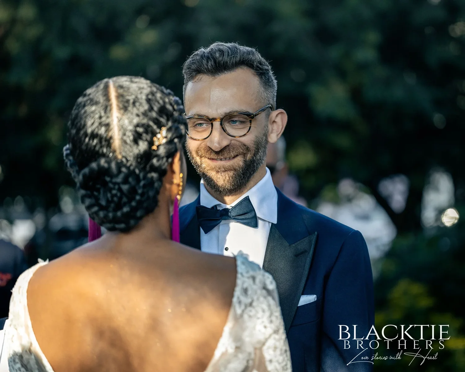 Drew smiling at Ciara during the vows, seen over her shoulder, his glasses and bow tie in sharp focus