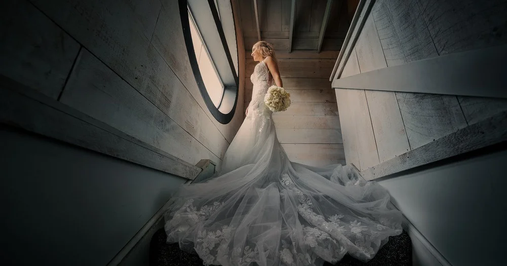 Jennifer in her wedding dress at the top of a whitewashed stairwell at The Cannery, lit by a round window, her train spread down the steps
