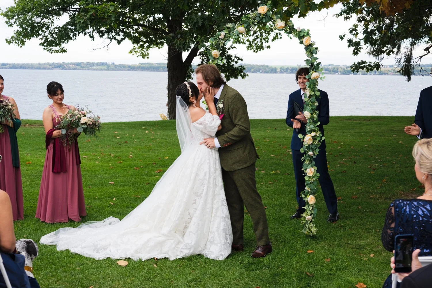 Marseille and Jason’s first kiss beside the rose arch, with Seneca Lake behind them and the wedding party looking on
