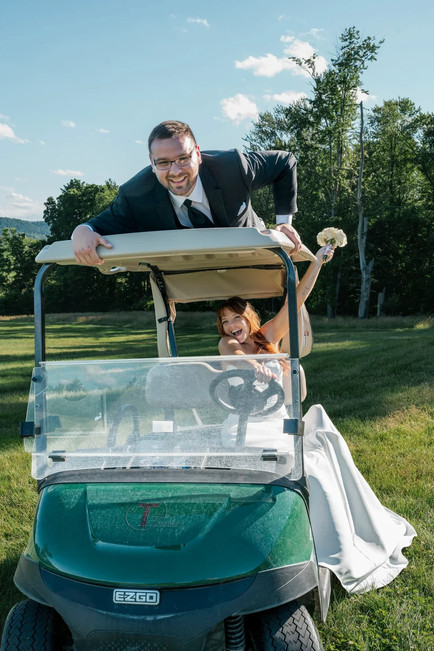 Zach leaning over the roof of a green golf cart while Danielle laughs in the driver's seat with her bouquet in the air, on the grounds at Traditions at the Glen
