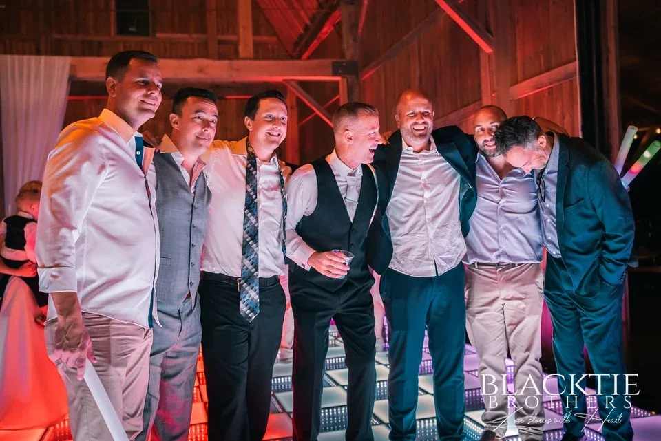 Guests arm in arm on the dance floor in the barn at Graceful Gatherings at LedgeRock, lit by coloured uplighting.