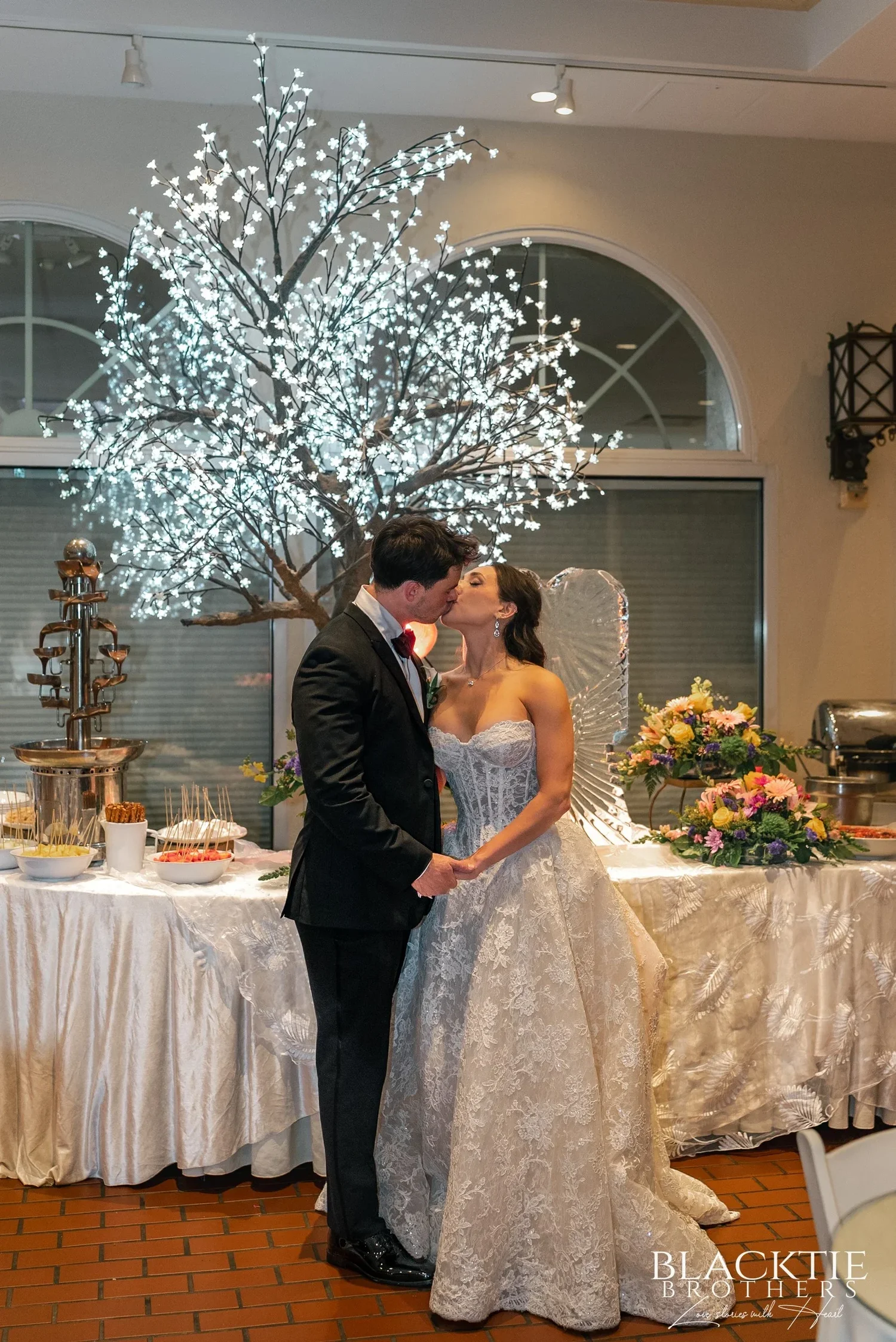 Leah and Nicholas kissing beside a tree lit with white lights at the dessert table, a chocolate fountain and trays of desserts next to them