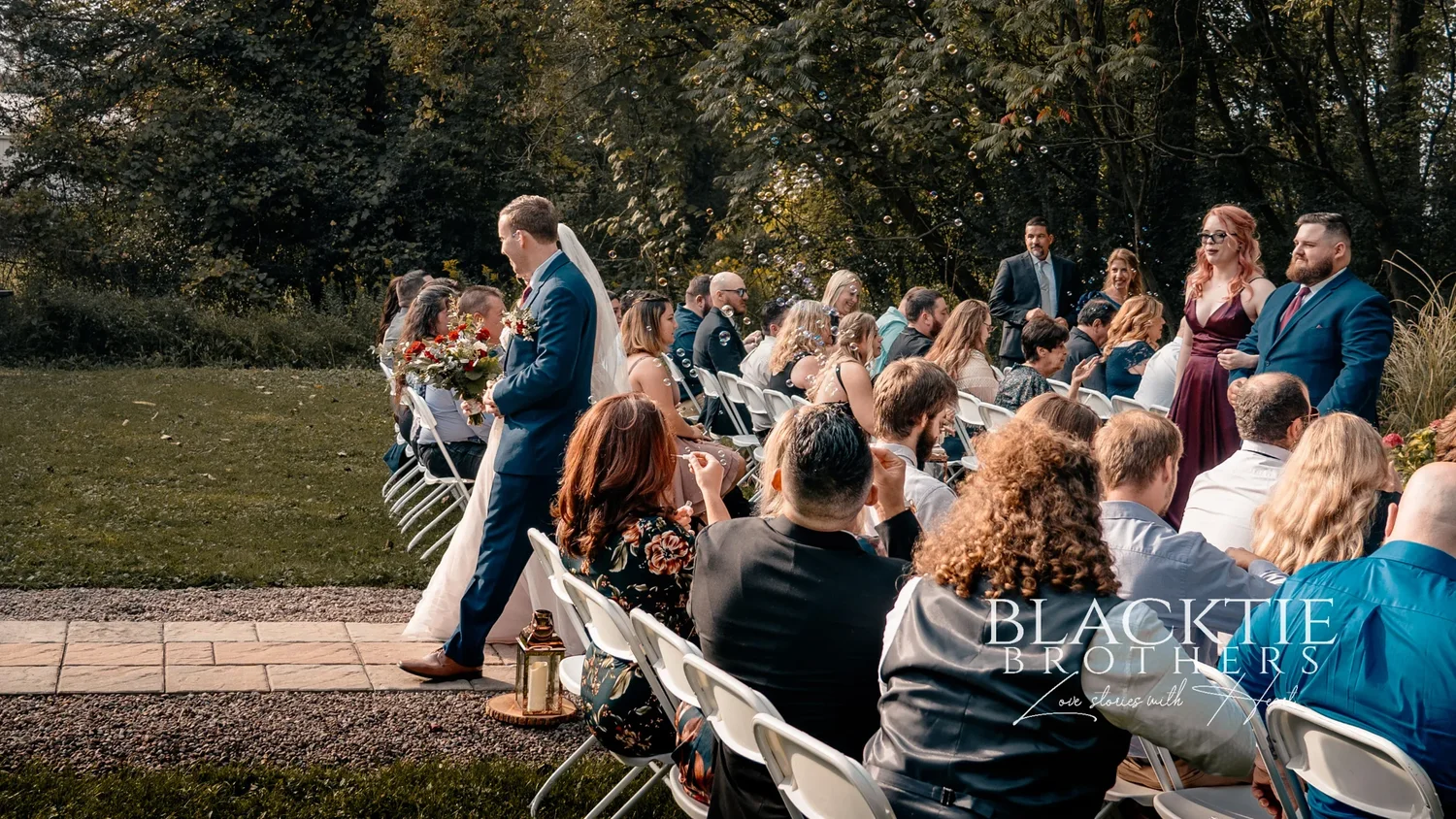 Jared walking down the aisle toward the white arbor while seated guests turn to watch, trees and a fence behind them