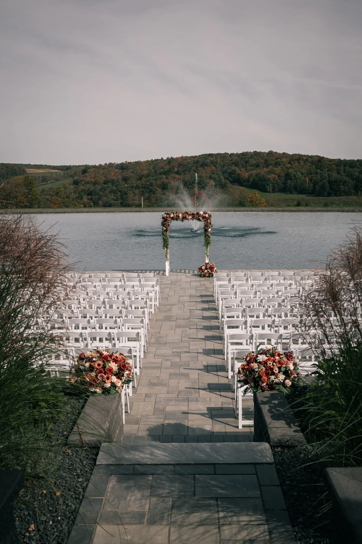 The empty ceremony aisle on the stone patio, white chairs and red flowers on either side, leading to the floral arch with the pond and hills beyond