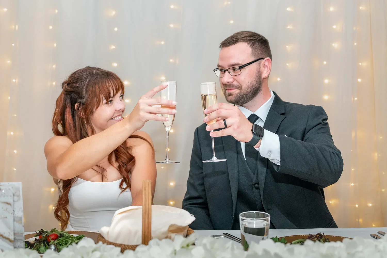 Danielle and Zach clinking champagne glasses at their sweetheart table in front of a curtain of string lights