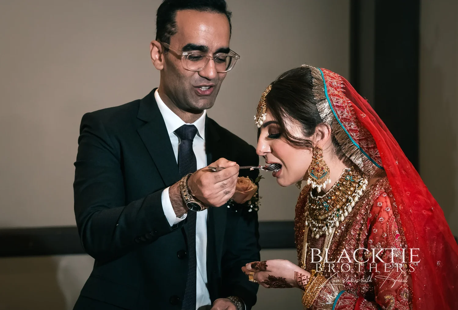 Rizwan feeding Jia a forkful of wedding cake in the ballroom, both leaning in over a small plate