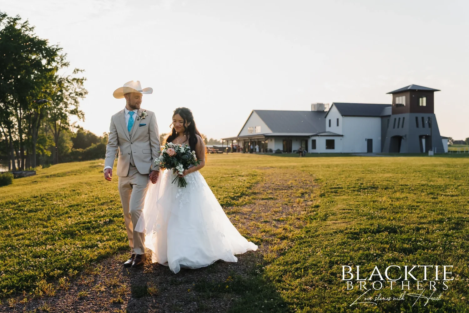 Carolina and Josh walking together across the lawn in the evening, Josh in a cowboy hat, the event center and its tower behind them