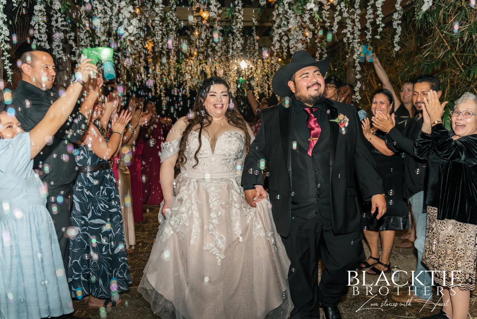 Alyssa and Alberto walk hand in hand through cheering guests and bubbles beneath hanging flowers and lights.