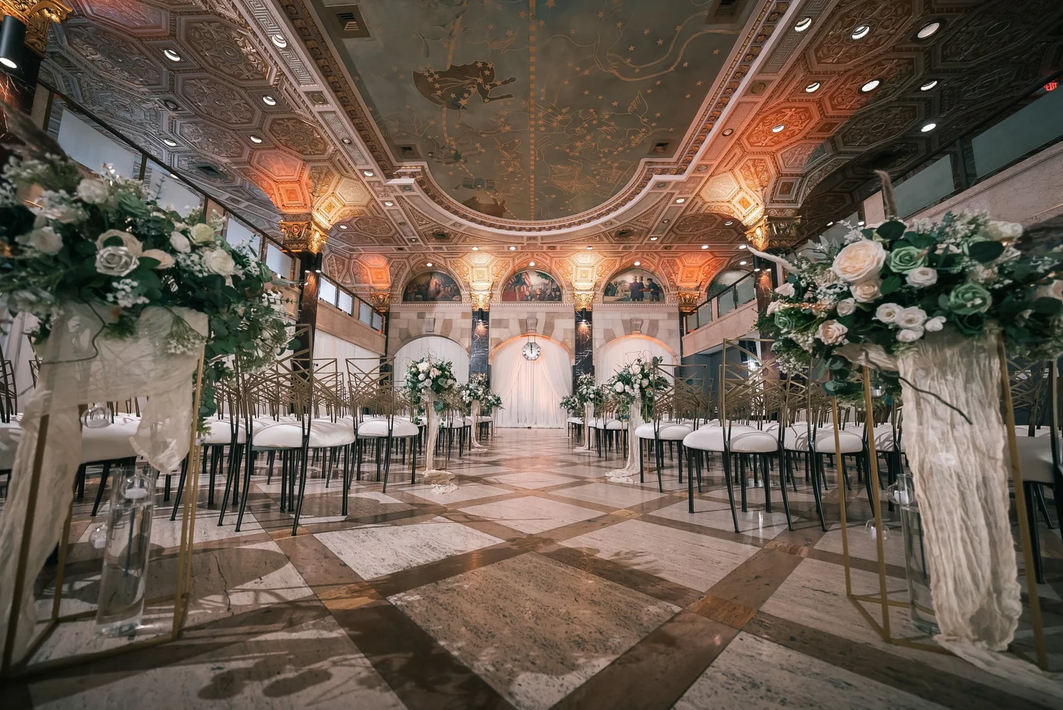 Rows of gold chairs with white cushions line a marble aisle toward a draped clock arch beneath Palladian Hall’s painted, coffered ceiling.