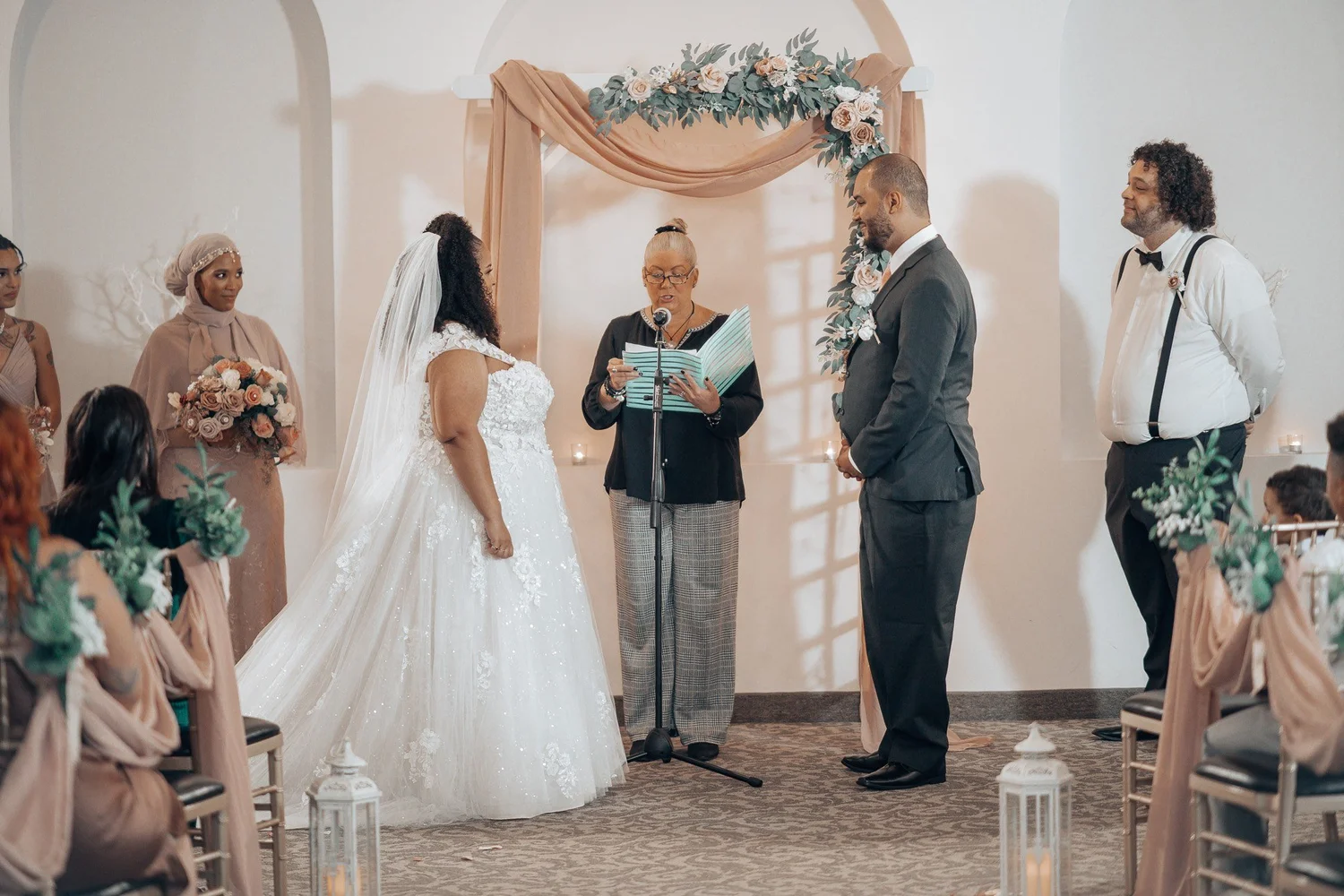 Christina and Erick facing their officiant under a floral arch draped in blush fabric, bridesmaids in blush dresses beside them
