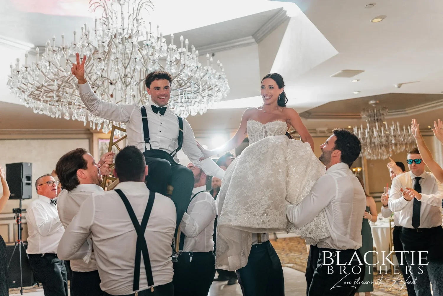 Leah and Nicholas lifted onto the shoulders of their friends on the dance floor, arms raised, under a crystal chandelier