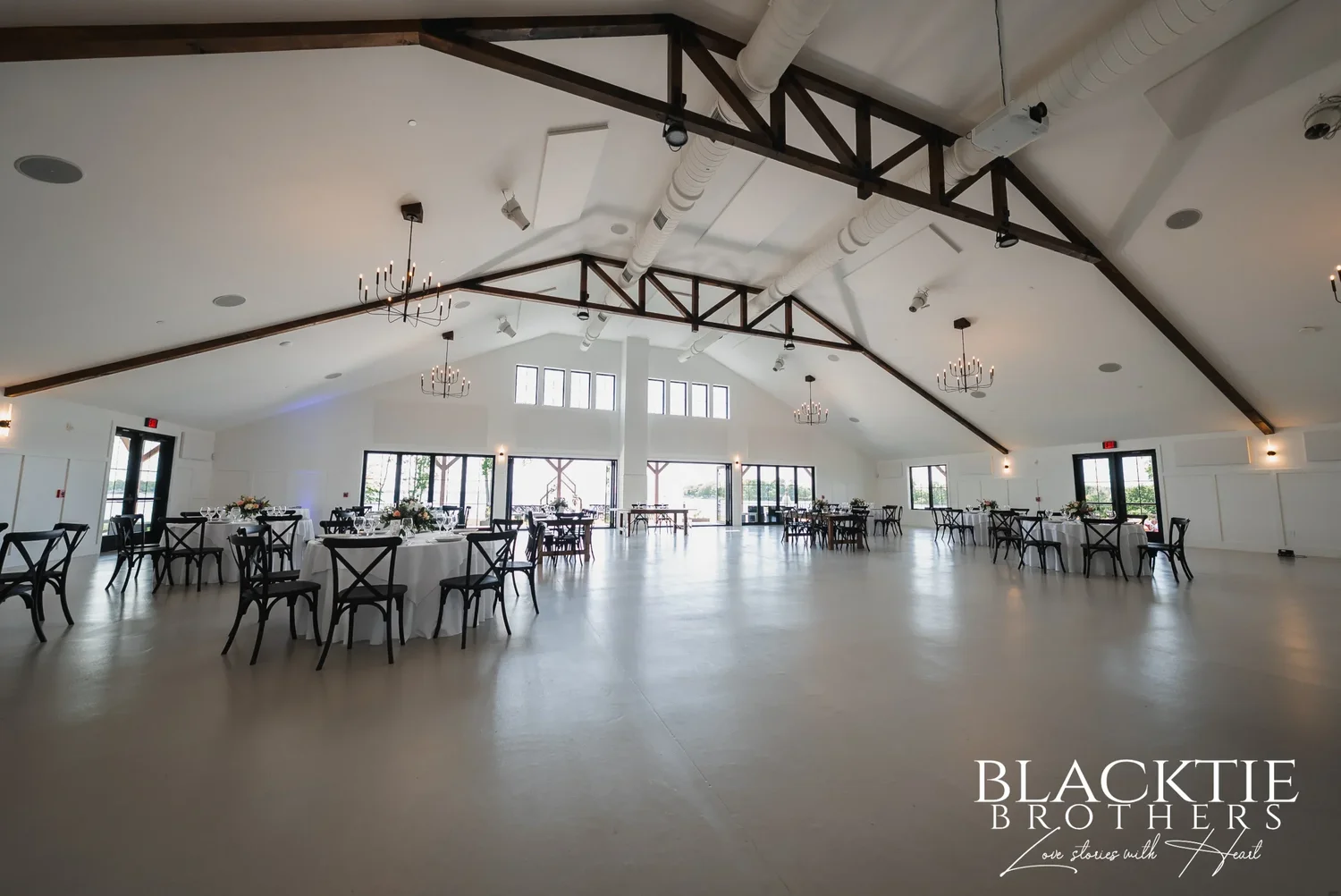The main hall at Sally Port View before guests arrive: white walls, dark timber trusses, chandeliers, round tables and a wall of black-framed glass doors at the far end