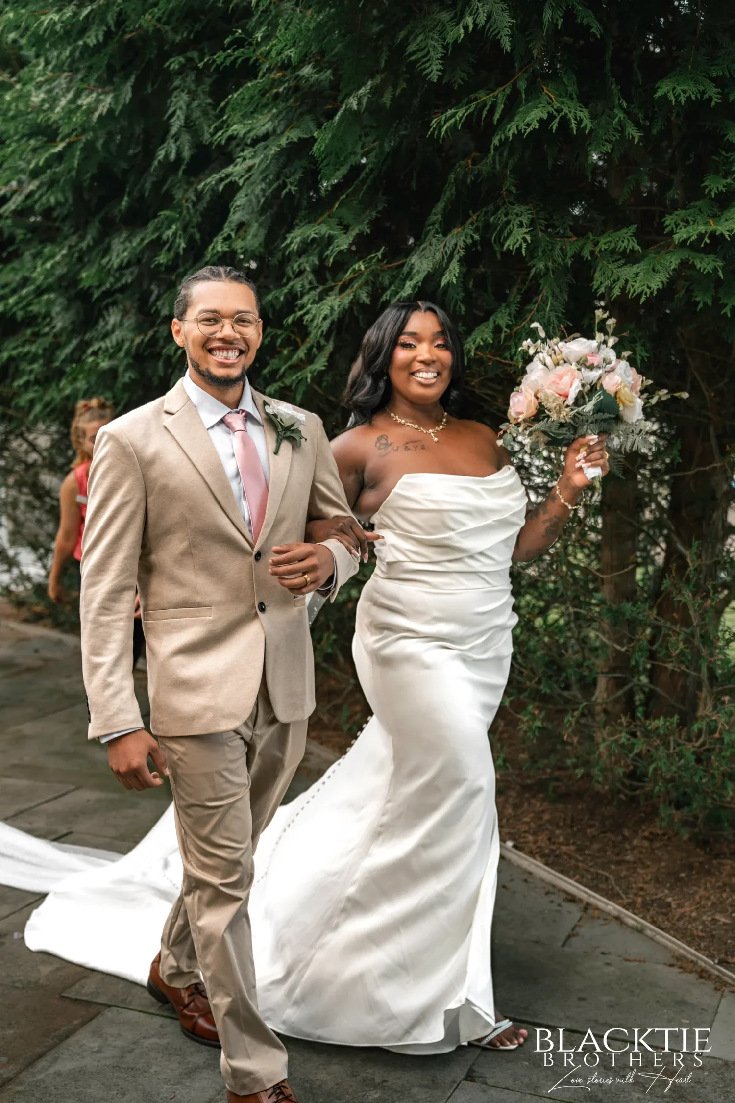 A groom in a tan suit and his bride, holding up her bouquet, walking back up the aisle after their ceremony at Traditions at the Glen