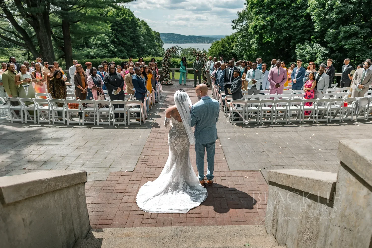 The couple at the top of the brick aisle facing their standing guests on the terrace, the Hudson River and hills in the distance
