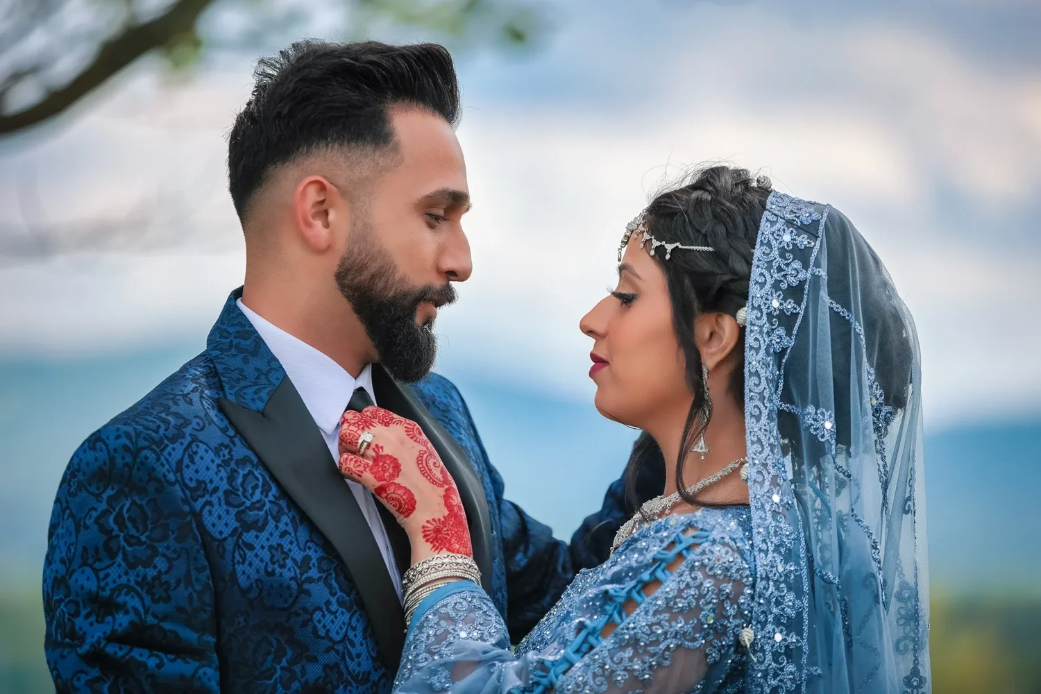 A bride resting her hennaed hand on her groom's lapel as they stand face to face on the hill at Traditions at the Glen
