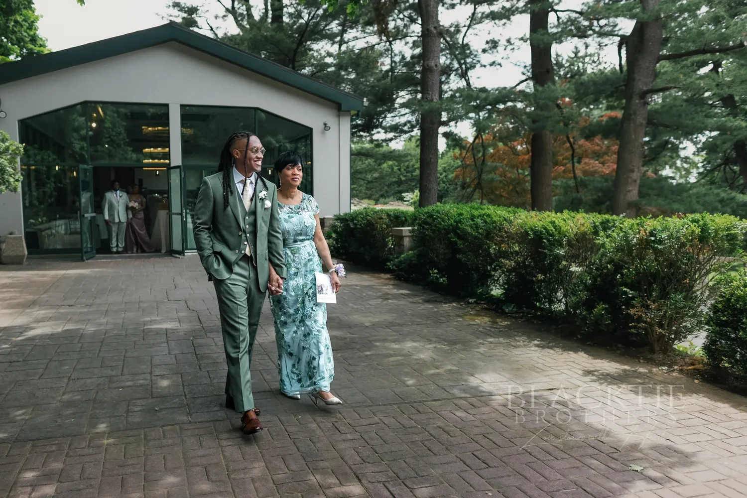 The groom walking across the terrace toward the ceremony beside a woman in a pale green dress, a white glass-walled building behind them