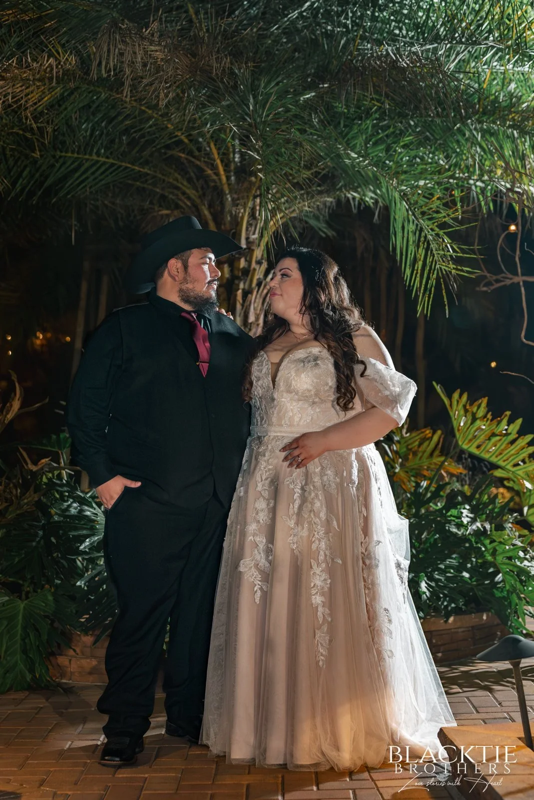 Alyssa and Alberto face each other beneath palm fronds in a garden portrait after dark.