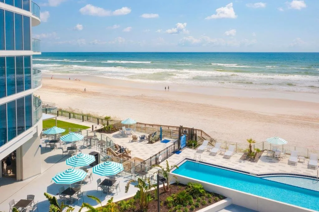 Max Beach Resort Daytona Beach aerial view of curved glass tower with oceanfront pool deck, blue-and-white striped umbrellas, and Atlantic beach