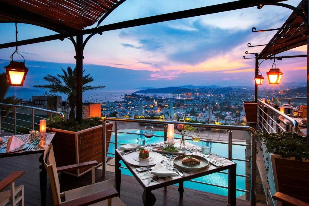 The Marmara Bodrum hilltop restaurant terrace at dusk with hanging lanterns overlooking Bodrum harbor and city lights below
