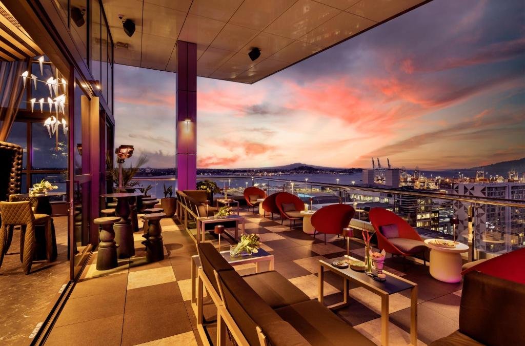 SO/ Auckland Auckland HI-SO rooftop bar terrace at sunset with red egg chairs, harbour water and port cranes visible under an orange and pink sky