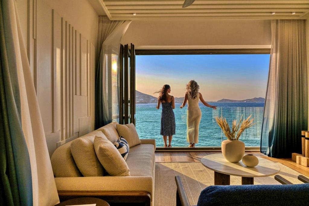 Cove Paros suite interior with bifold glass doors open to Aegean Sea and island silhouette at sunset