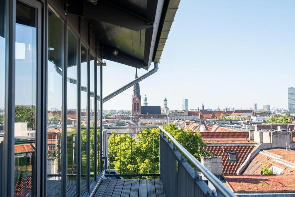 sly Berlin Berlin upper-floor balcony with glass sliding doors overlooking Friedrichshain rooftops with a red-brick Neo-Gothic church spire and distant city towers on the horizon