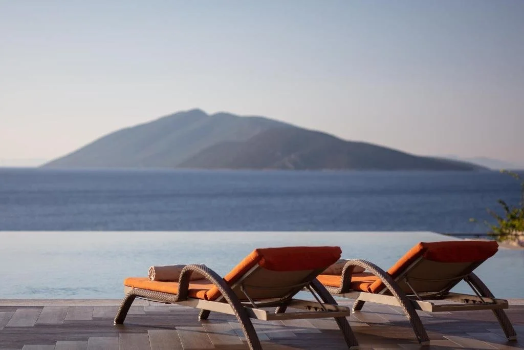 Caresse Luxury Collection Bodrum infinity pool terrace with orange-cushioned sun loungers and island silhouette on the Aegean horizon