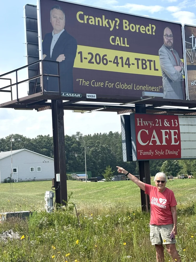 Photo of Hariet in shorts and a red t-shirt standing and pointing under the TBTL billboard, which is a parody of a lawyer's billboard that says "Cranky? Bored? Call 1 206-414-TBTL"