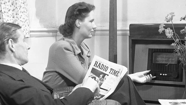 Black and white photo apparently from the2940 of a man and woman in a living room. The woman is turning a dial on a big living room radio, while the man is reading a newspaper called RADIO TIME.
