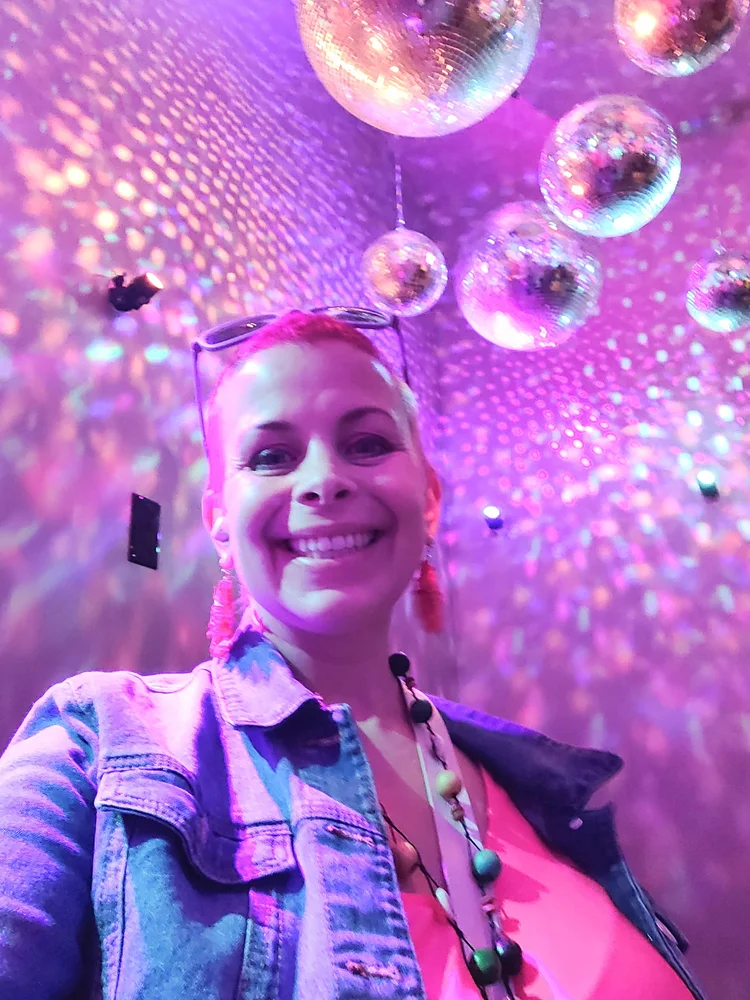 Photo of listener Kelley in a pink-lit room with a bunch of disco balls. Kelley is smiling big for the selfie. 