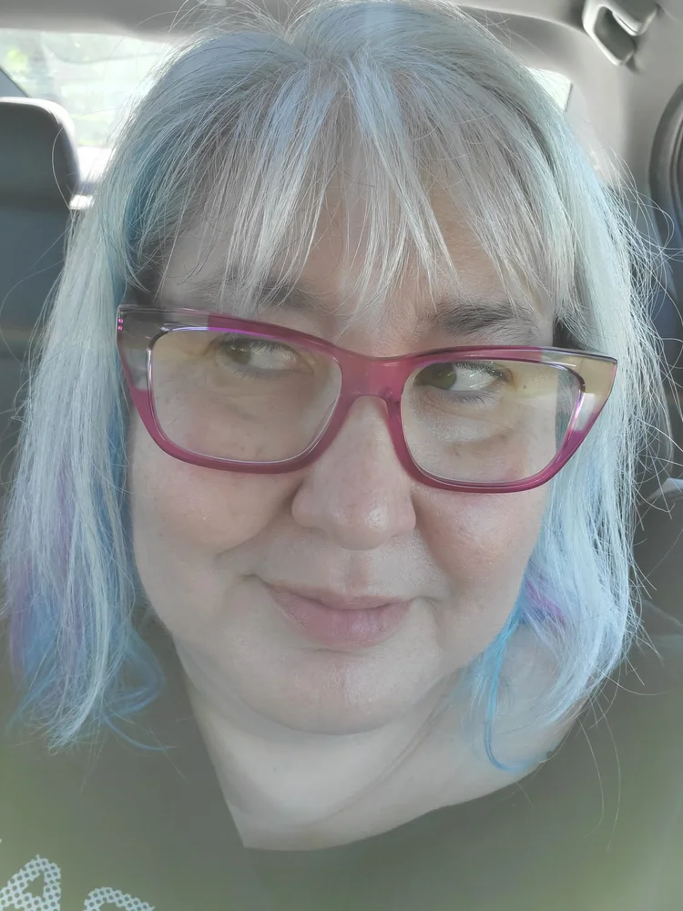 Photo of Listener Elissa, I believe sitting in a car, closeup on her face, wearing red chunky glasses