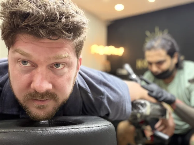 Photo of listener Max laying down on a tattoo table.  His face is in focus right close to the camera, and we see a tattoo artist in the background working on a tattoo on Max's arm or leg.  