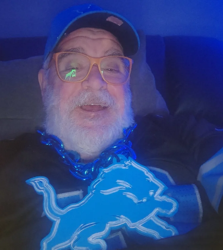 Photo of listener Ron, leaning back on a couch in a mostly darkened room with blue light from a TV. He's got cool, biggish orange glasses, a white beard, a Detroit Lions hat and a huge Lions plastic pendant around his neck.