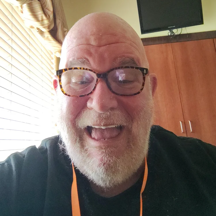 Photo of listener Ron. Looks like a selfie next to a window with miniblinds. He's bald with a short white beard and glasses. He's smiling into the camera very nicely! 