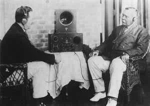 Very old photo of two men in front of either a recording device or listening device. One guy looks a lot like Teddy Roosevelt.