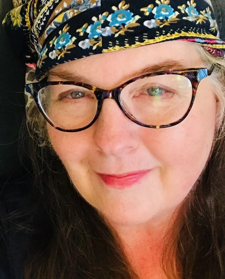 Photo of listener April. Sher's wearing a colorful headscarf or cloth hat and brown glasses. It's a closeup portrait. 