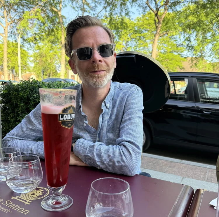 Photo of listener Matt sitting at an outdoor cafe with a tall, red drink in front of him. He's wearing sunglasses and half-smiling at the camera