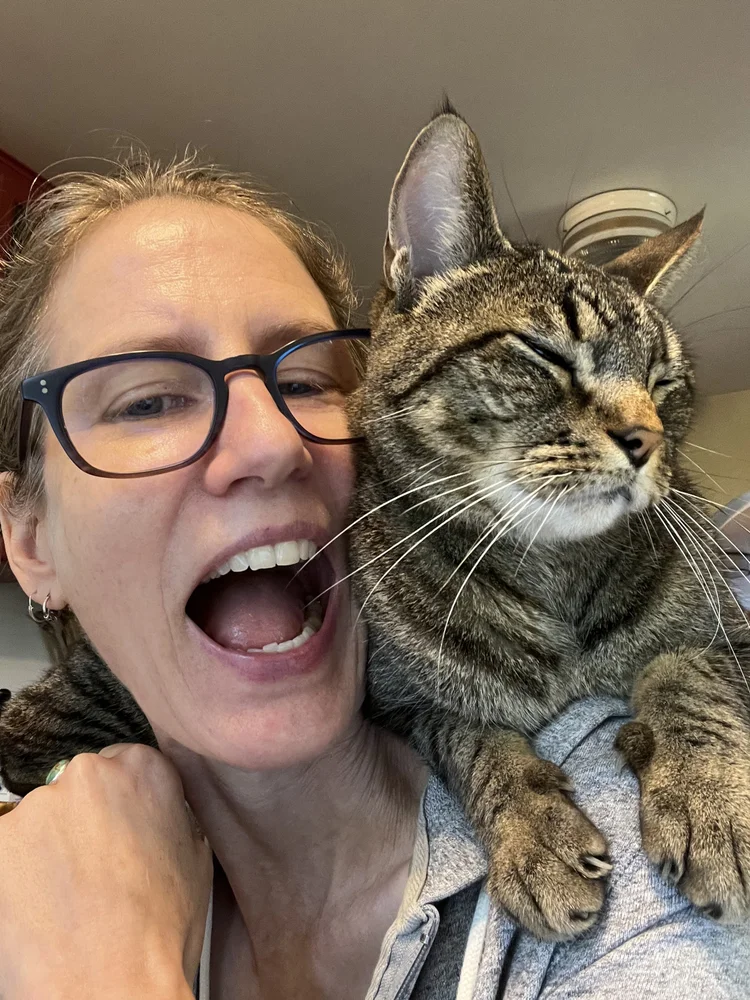 Photo of Listener kim with a cat on her shoulder! Kim has her mouth open in a wide smile and the cat is squinting in happiness