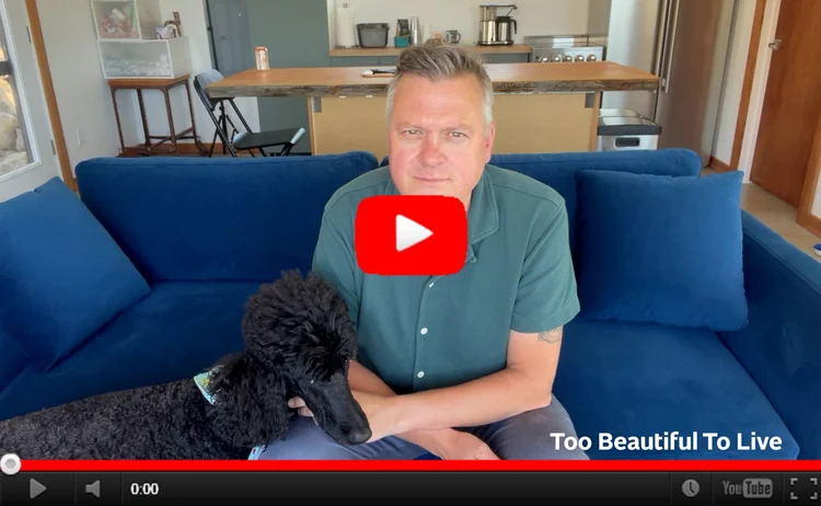 YouTube still image of Luke sitting on a blue couch petting a black standard poodle standing next to him on the floor