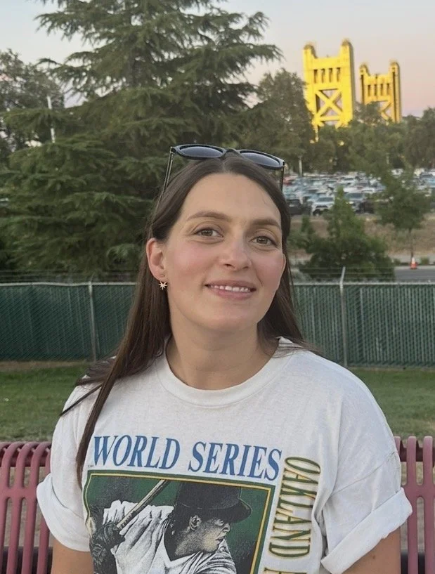 Photo of listener Kelsey sitting on a bench outside with sunglasses  pushed up on top of her head and a kickass Oakland As World Series shirt.