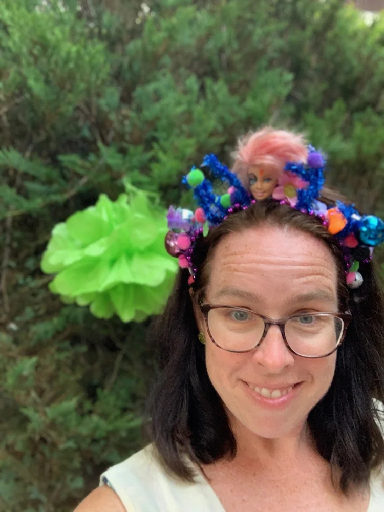 Photo of listener Cristin outside somewhere with a very involved headdress on that appears to include a big green flowery thing and the head of a Barbie doll? 